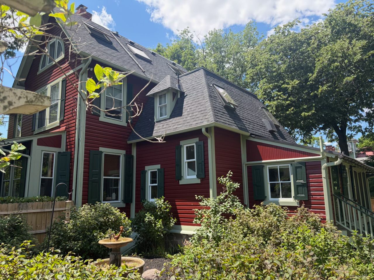 Red house with steep gray roof and white trim, surrounded by trees and shrubs on a sunny day