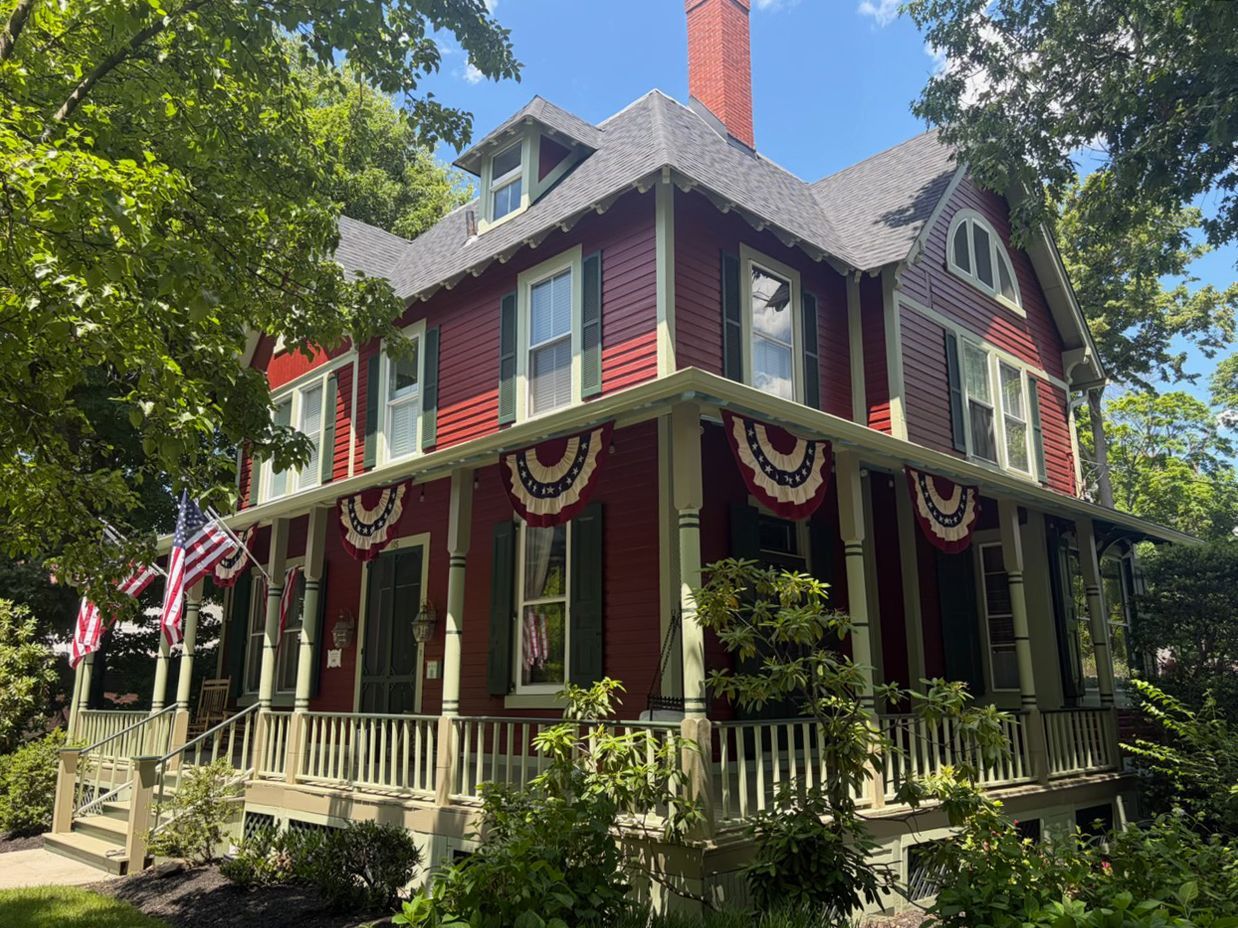 Victorian house with red siding, white trim, and patriotic bunting on a sunny tree-lined street