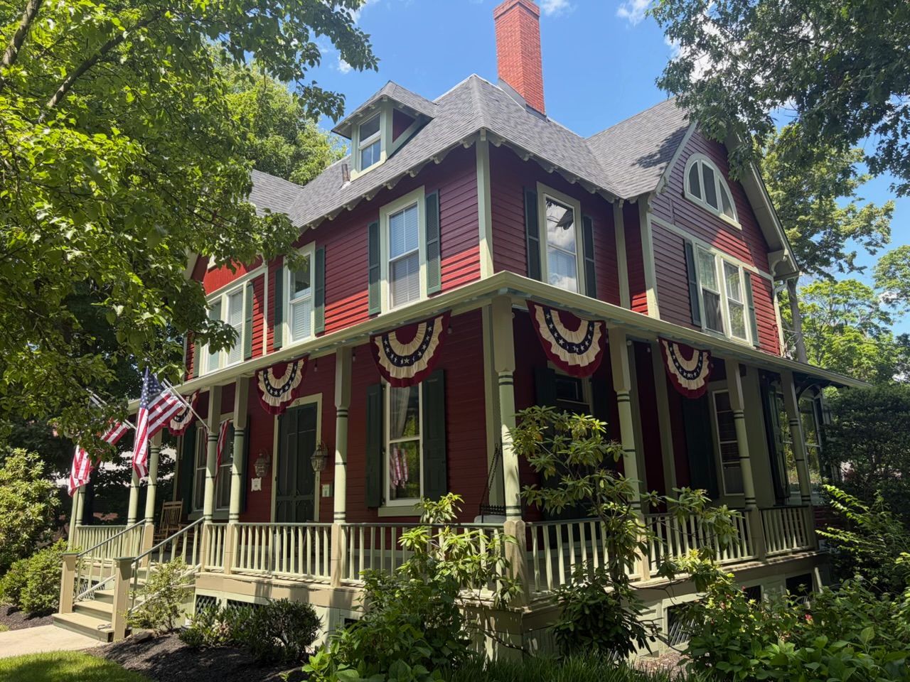 Victorian house with red siding, white trim, and patriotic bunting on a sunny tree-lined street