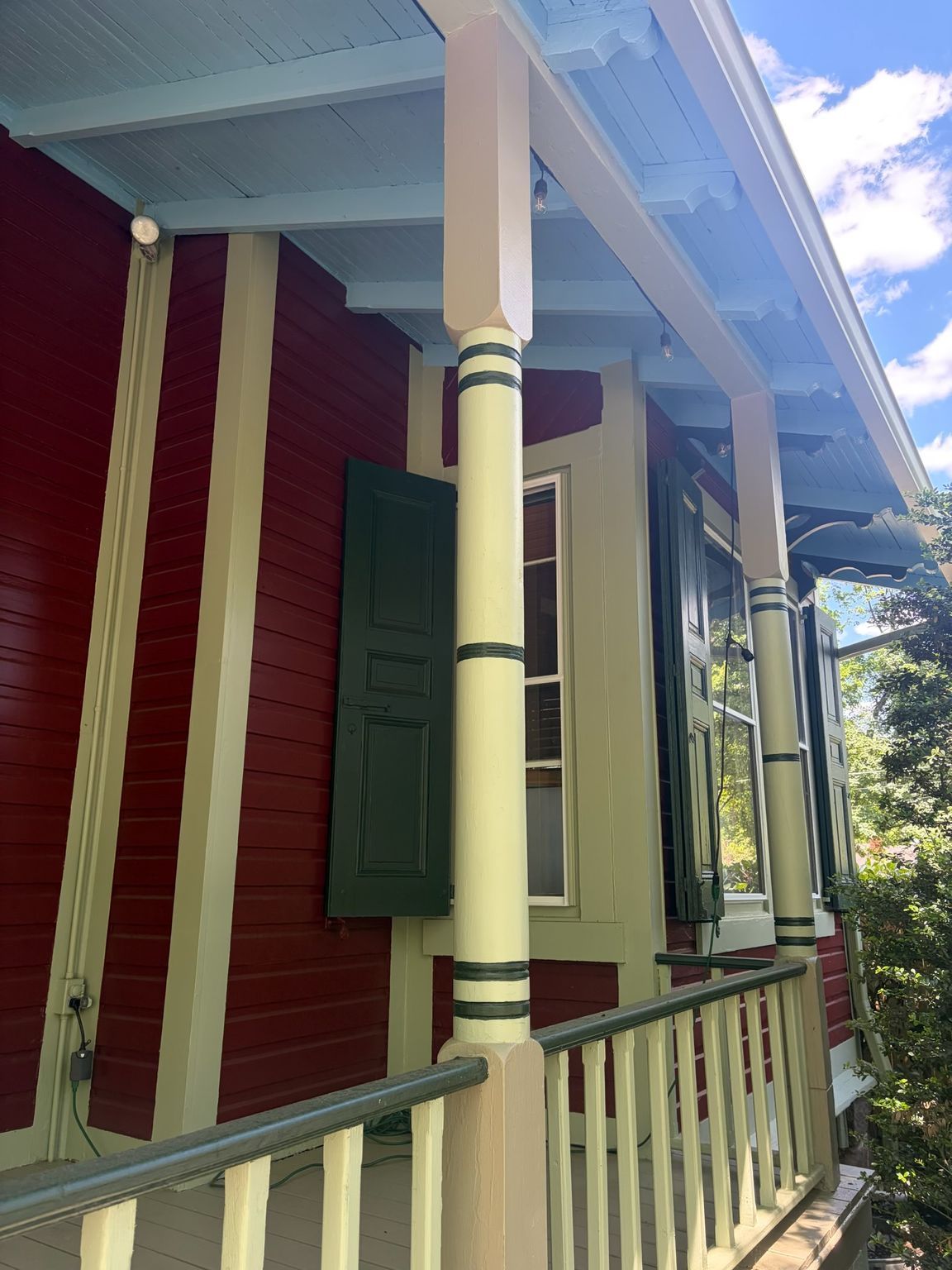 Red house porch with green shutters, cream trim, and white railing in daylight.
