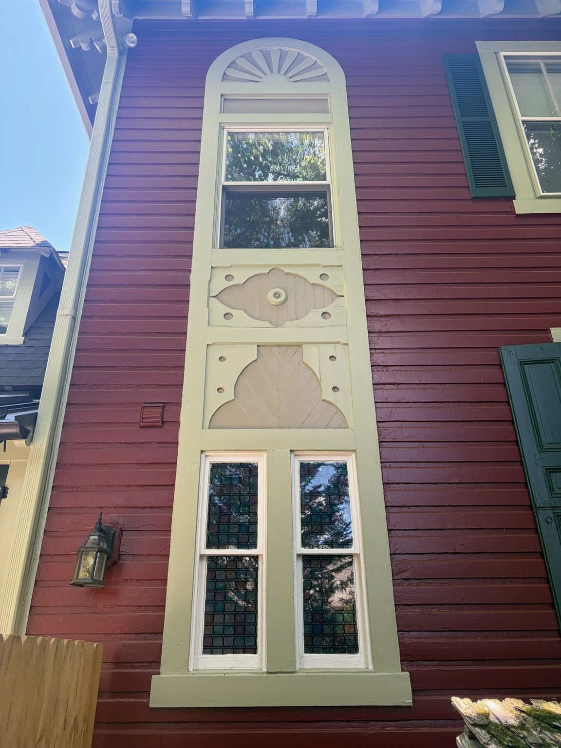 A tall, narrow decorative window set in a red house wall with cream trim and green shutters