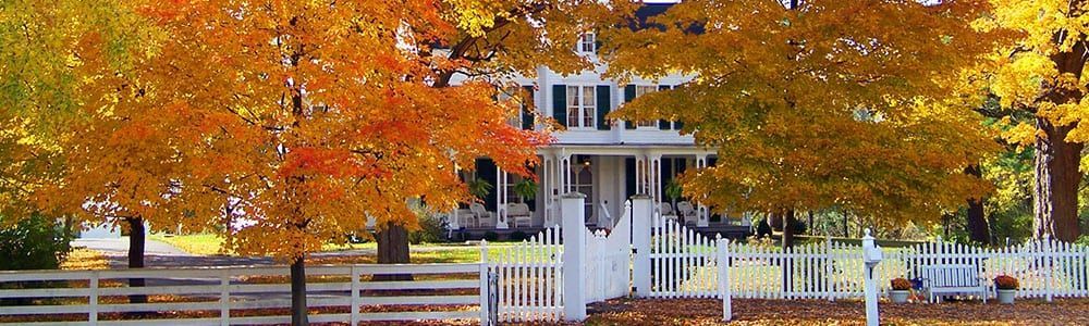 Autumn trees behind a white fence and house, with bright orange leaves