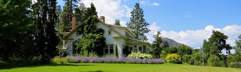 White farmhouse with porch amid green lawn, flowers, and tall trees under a bright blue sky.