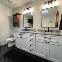 White bathroom with double sinks, mirrors, vanity cabinets, and a toilet beside a dark floor rug