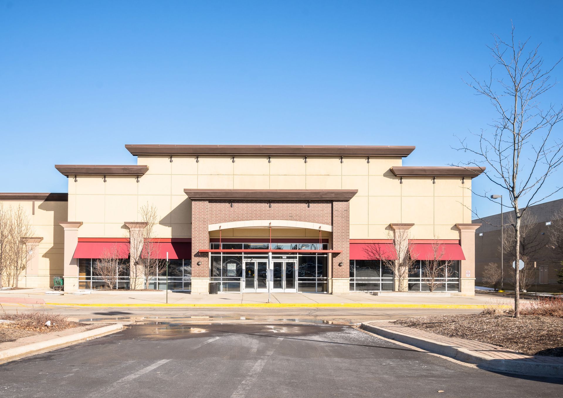 Exterior of a vacant, beige commercial building with red awnings and a brick entrance, under a clear blue sky.