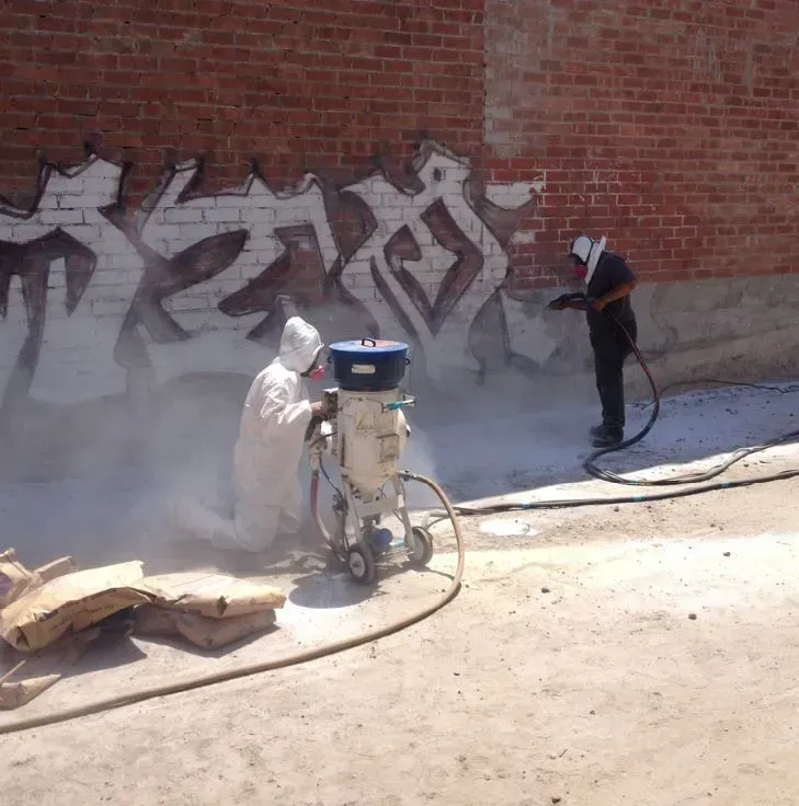 Two workers removing graffiti from a brick wall with sandblasting equipment, creating a cloud of dust.