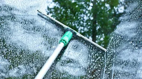 A window being cleaned with a squeegee, soapy water visible; green handle, white pole.
