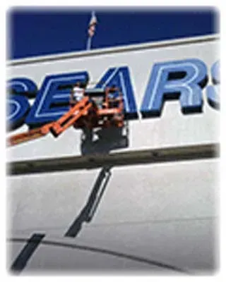 Maintenance worker in lift repairing Sears sign on a white building, blue and white letters.