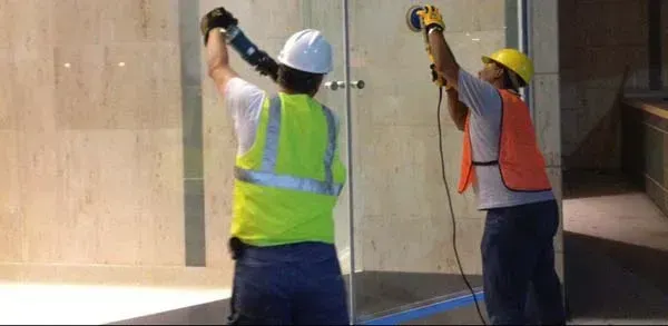 Two construction workers in safety vests are working on glass. One holds a level, the other uses a power tool.