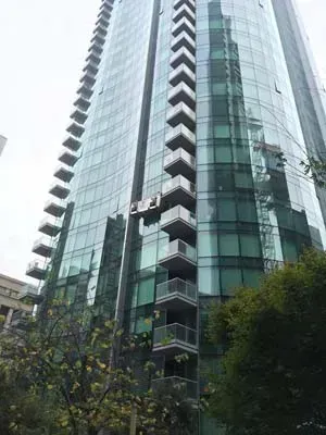 Tall glass high-rise with balconies. Green-tinted glass reflects the sky and trees below.