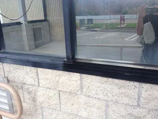 Window with black trim on a beige brick wall reflecting a parking lot and person.