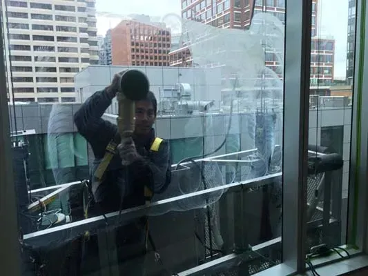 Person using a tool to work on the exterior of a window. City buildings are in the background.