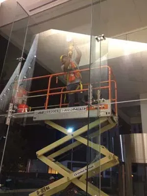 Two workers on a yellow scissor lift installing glass panels on a building at night.