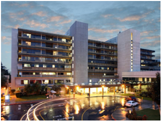 Hospital building exterior at dusk, with lights on, reflecting in a wet driveway.
