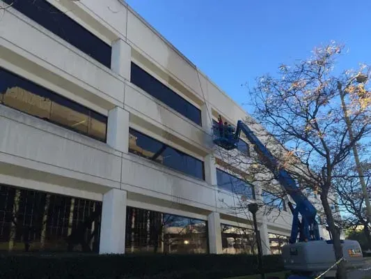 Construction workers in a lift, cleaning or repairing a beige building's exterior. Blue sky, leafless tree.