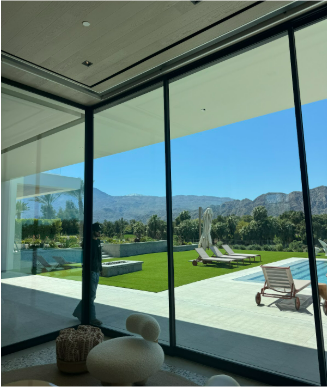 Large glass windows frame a view of a lush green lawn, pool, and mountains under a blue sky.