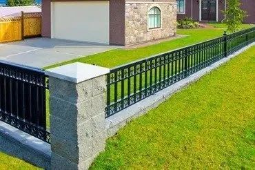 Black metal fence with stone pillars bordering a green lawn beside a house and driveway