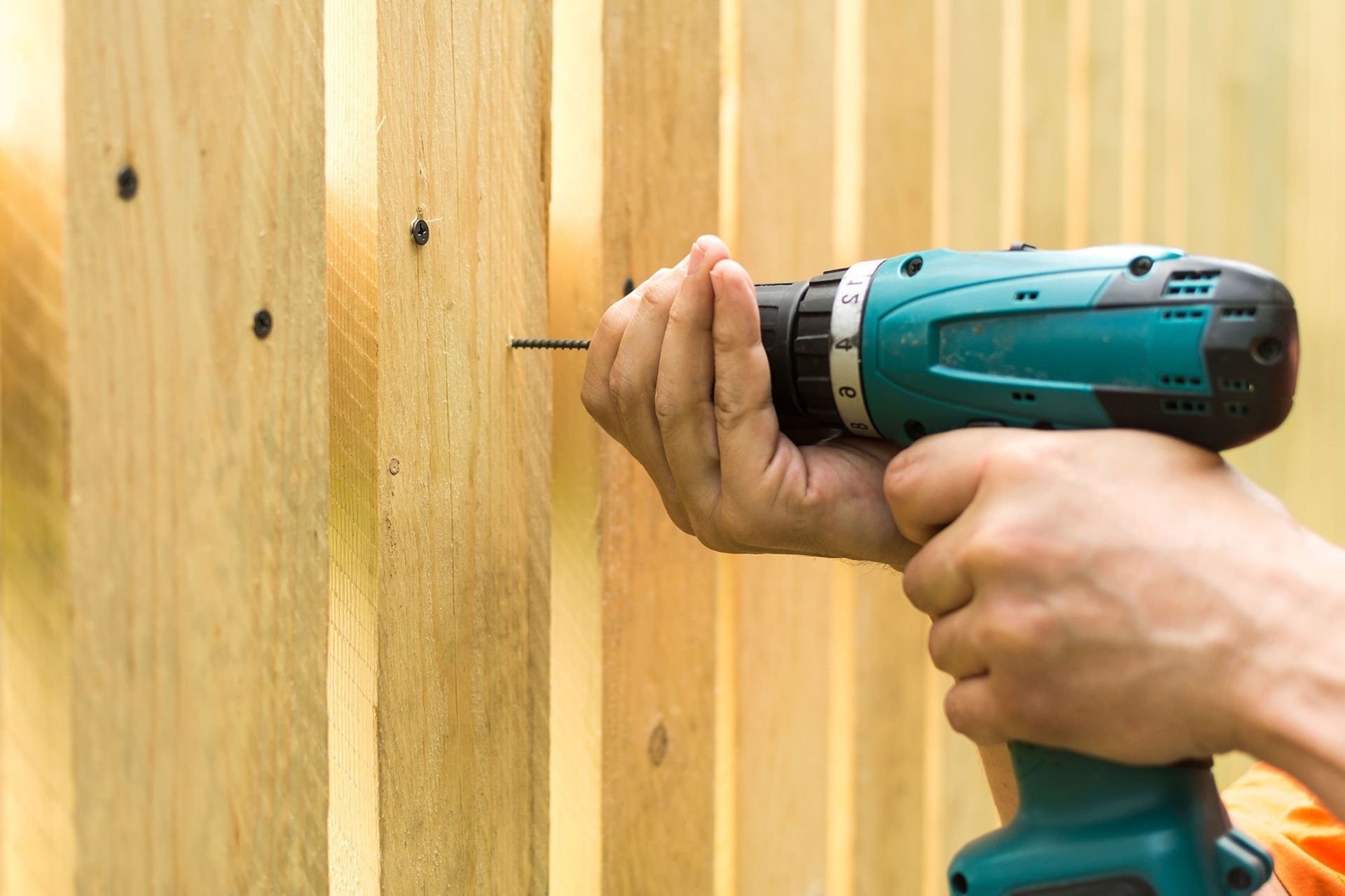 Hands using a teal cordless drill to drill into a wooden fence board.
