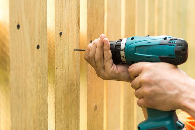 Hands using a teal cordless drill to drill into a wooden fence board.