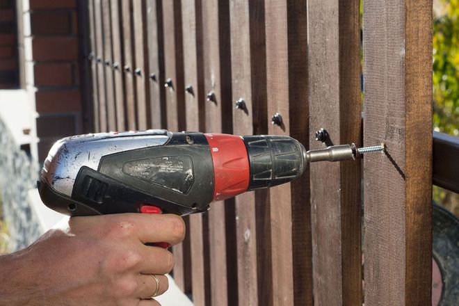 Hand holding a cordless drill driving a screw into a wooden fence panel