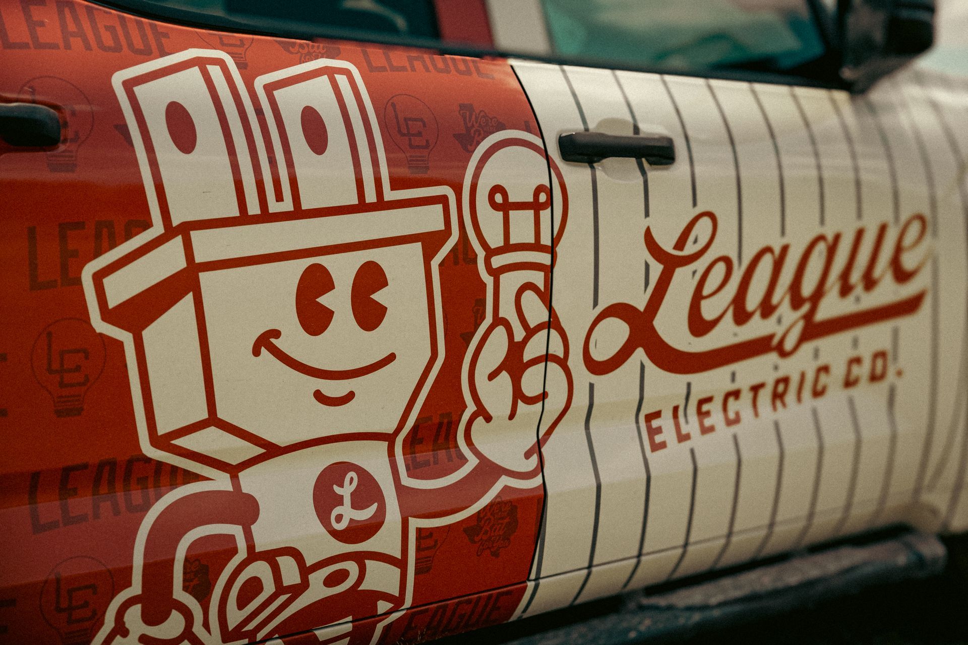 Red and white baseball-themed truck door graphic with a smiling plug mascot holding a lightbulb for "League Electric Co."