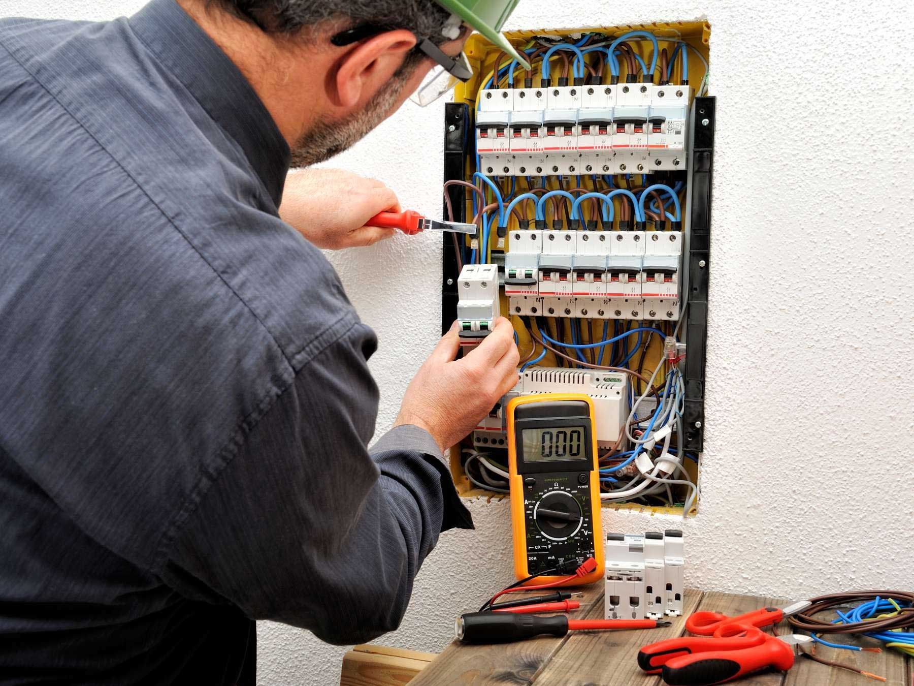 Electrician using a screwdriver and multimeter on a circuit breaker panel in a white-walled setting.