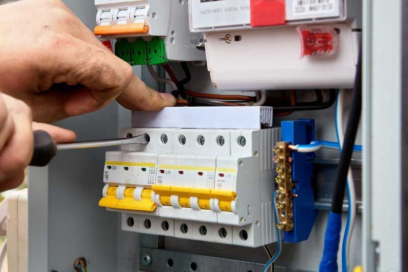 Person using a screwdriver to work on an electrical panel with multiple circuit breakers, wires, and terminals.