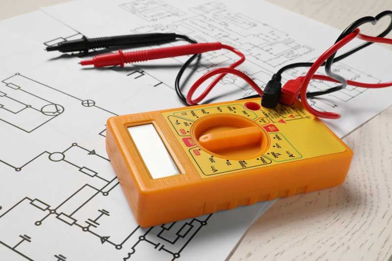 A yellow multimeter with attached red and black wires and probes sits on top of a circuit diagram.