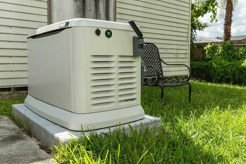 A beige generator on a concrete pad in a grassy yard next to a white house. A black metal chair sits behind it.