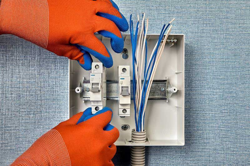 Hands in orange and blue gloves working on electrical wiring in a breaker box on a blue textured wall.