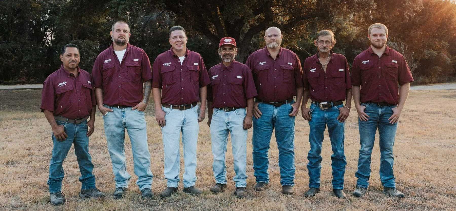 Seven men in maroon shirts and jeans standing outdoors, smiling.