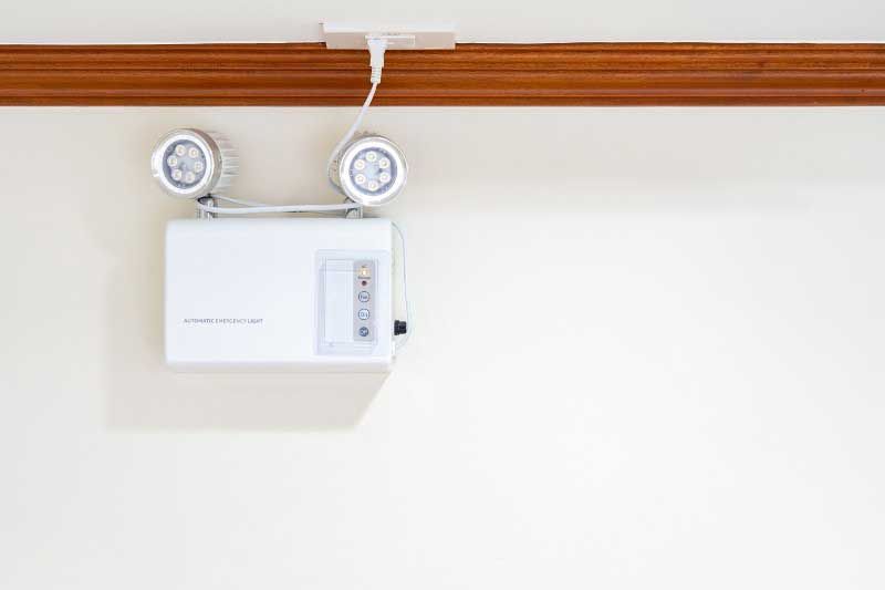 White emergency light fixture mounted on a white wall beneath a wooden trim. It has two spotlight heads.