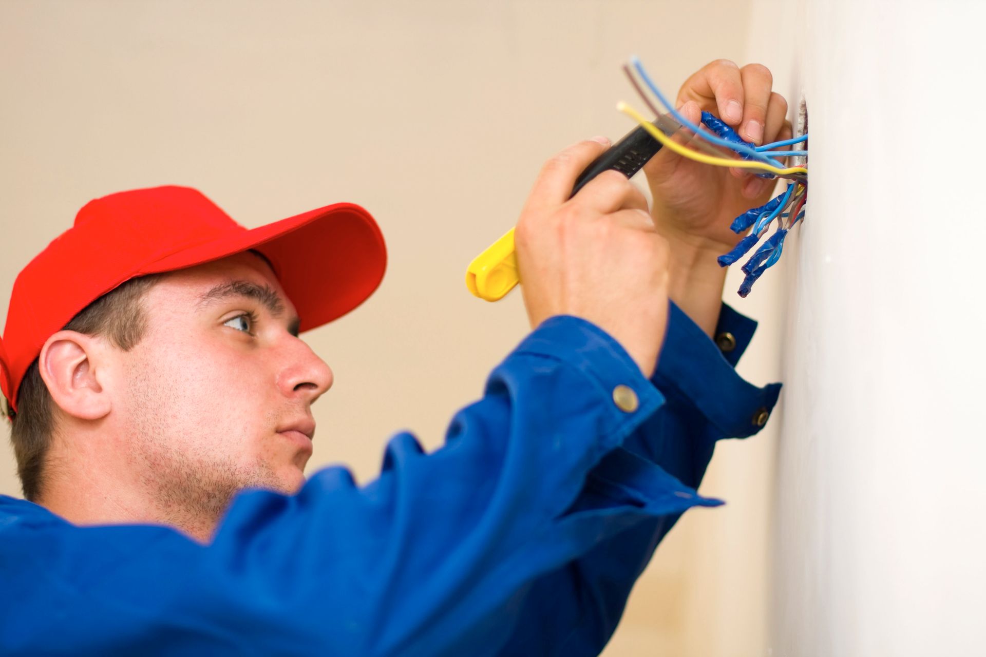Electrician in blue work clothes and a red cap wiring electrical cables into a wall outlet.