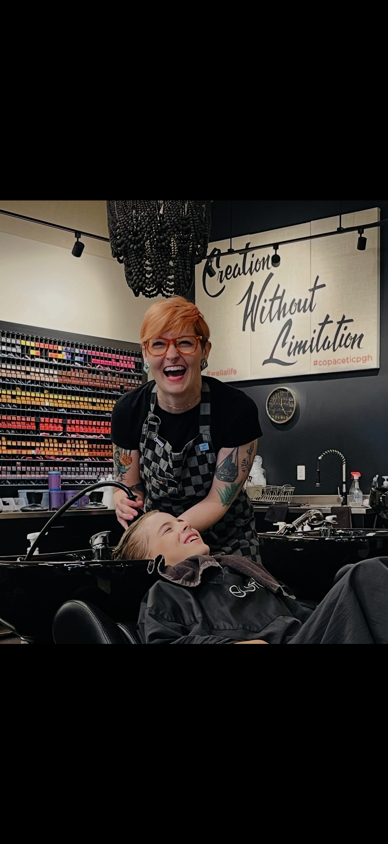 Young child with a fresh haircut holds a jar and smiles at a laughing woman in a salon setting.