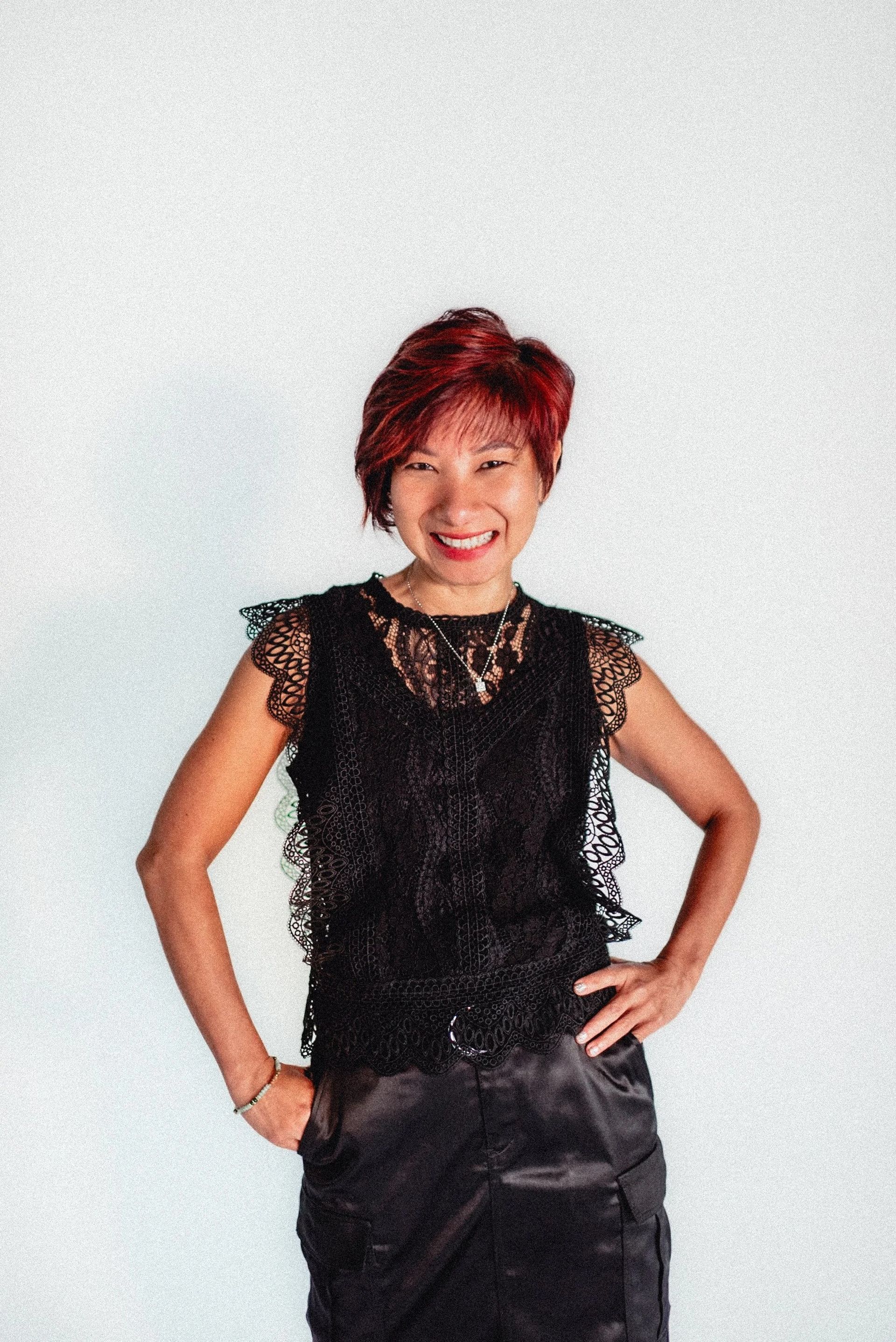 Woman with red hair, black lace top, hands on hips, smiling, against a white background.