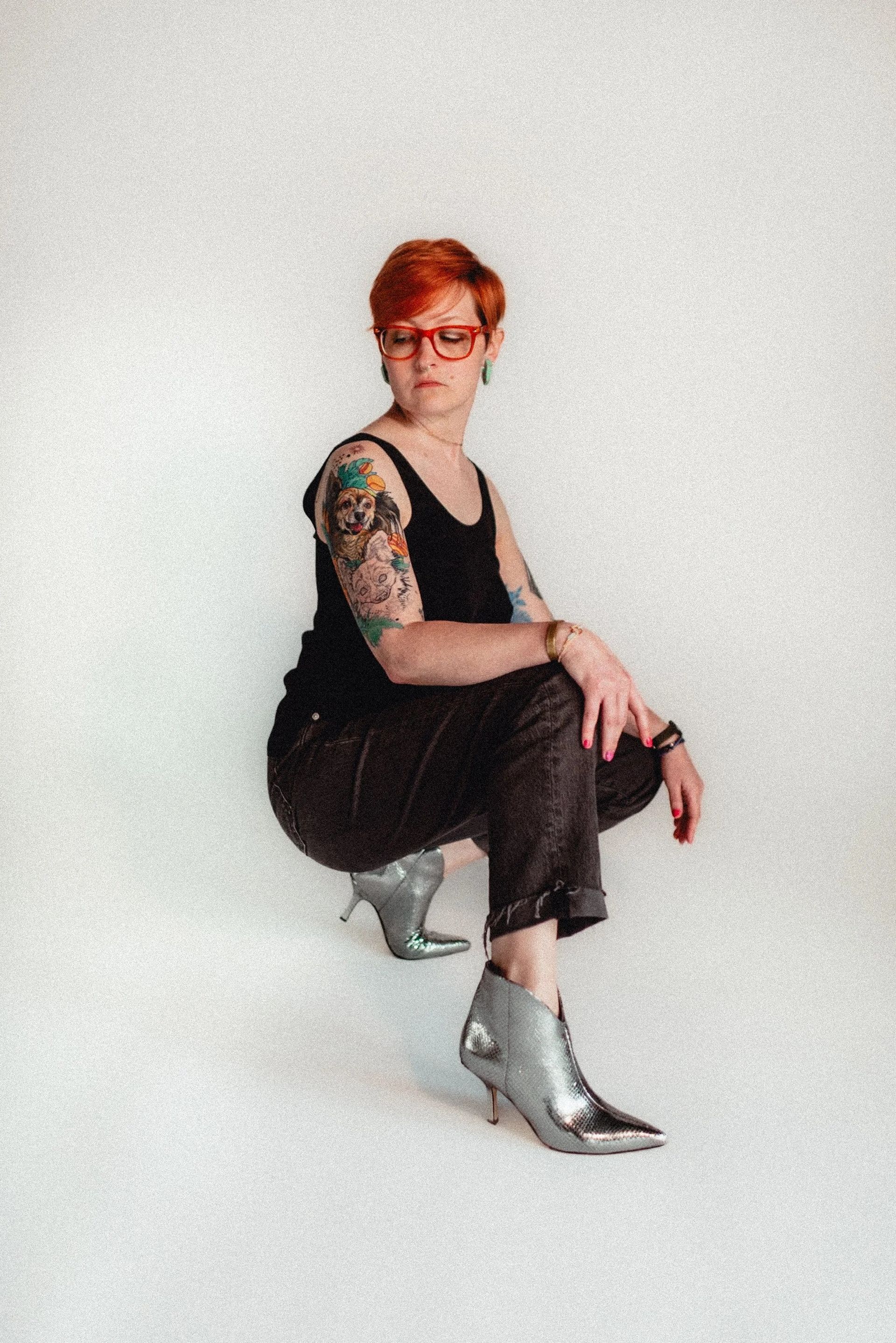 Woman with red hair, glasses, and arm tattoo squats, wearing a black top, pants, and silver high-heeled boots.