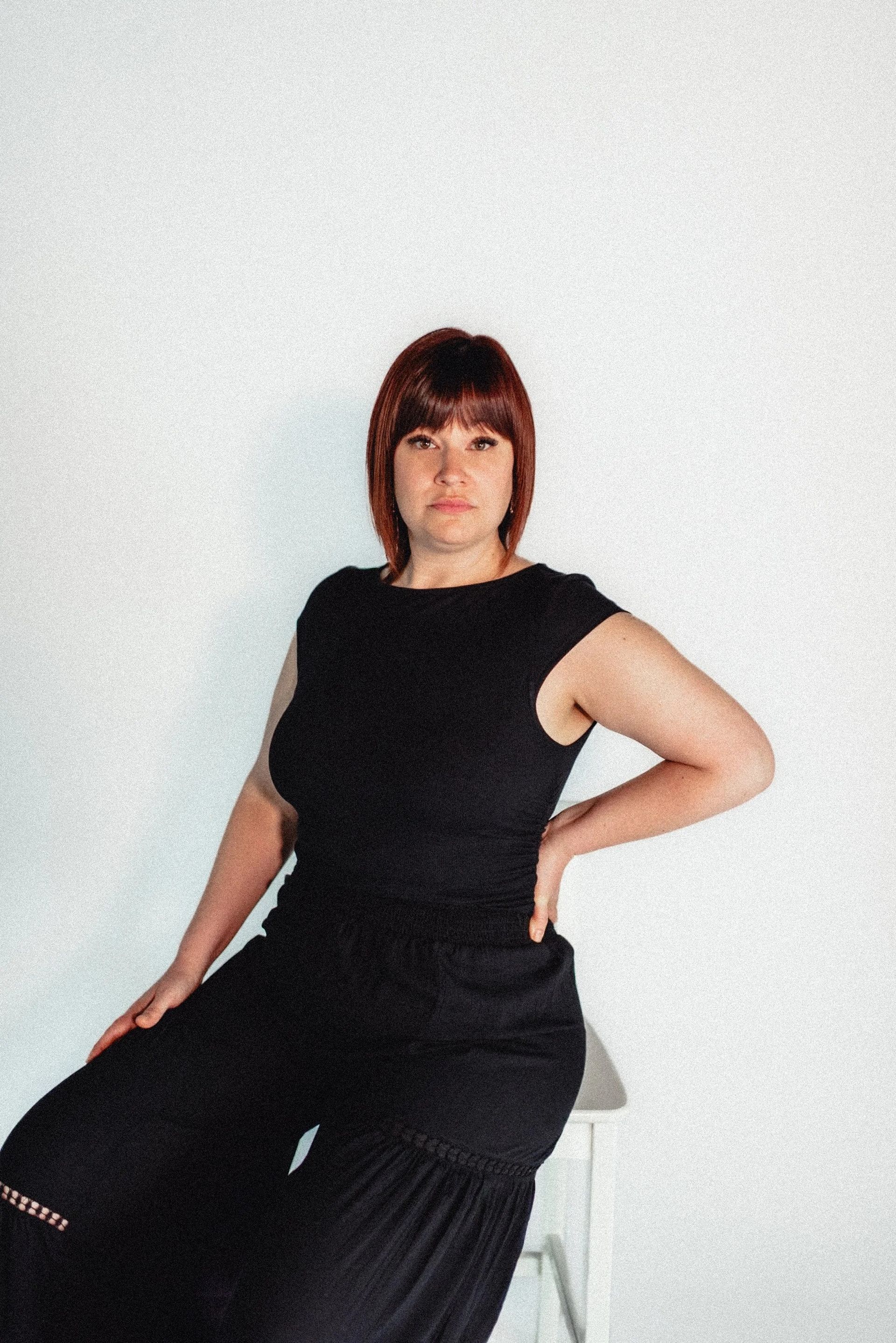 Woman in black outfit sits on a white stool, hand on hip, looking at camera against a white backdrop.