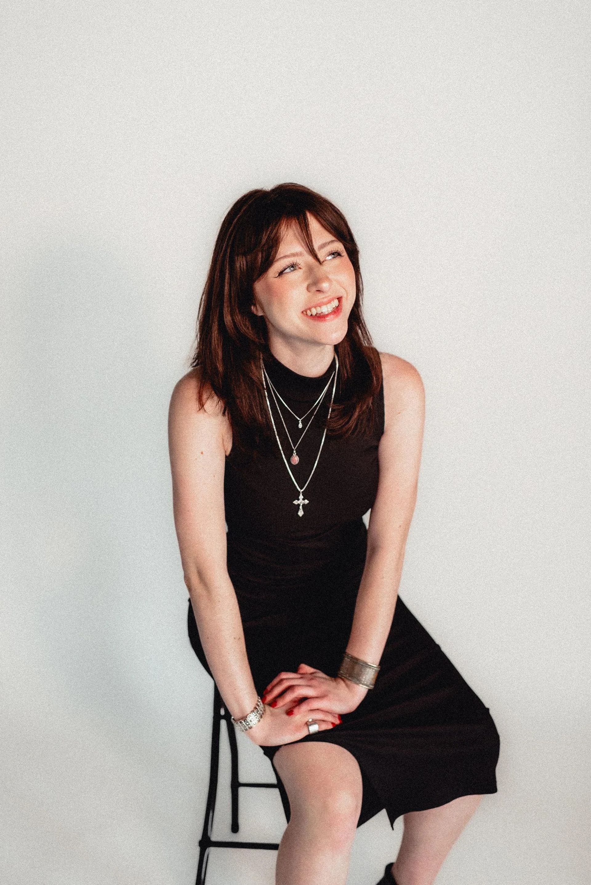 Woman in black dress, smiling, seated on a stool, looking upwards against white backdrop.