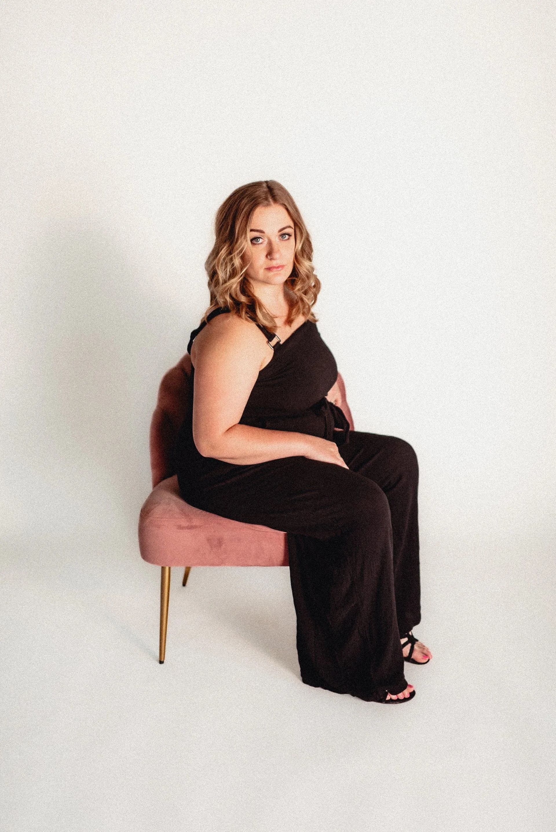 Woman in black jumpsuit sits on pink chair, looking at camera. White backdrop.