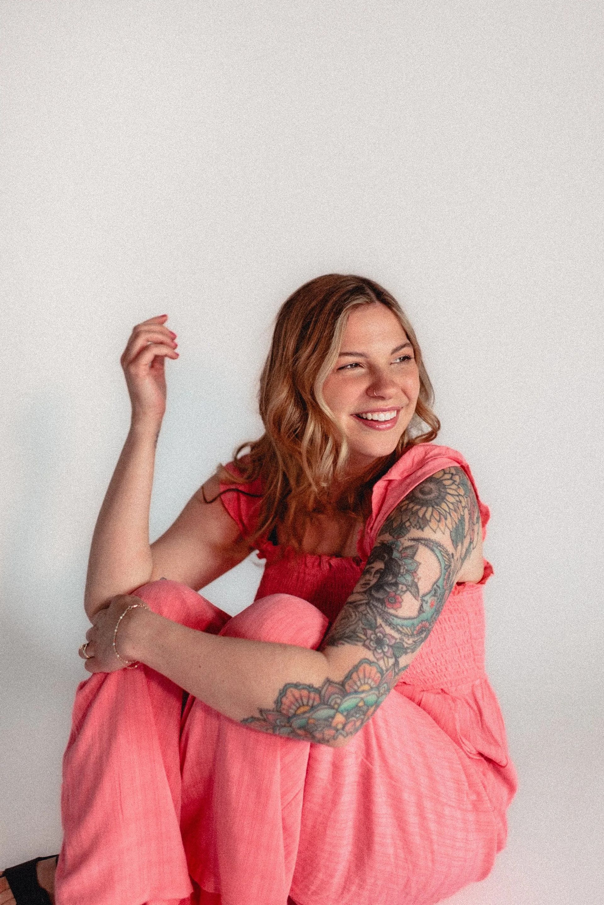 Woman with arm tattoos smiles, sitting cross-legged, wearing a coral jumpsuit, against a white background.