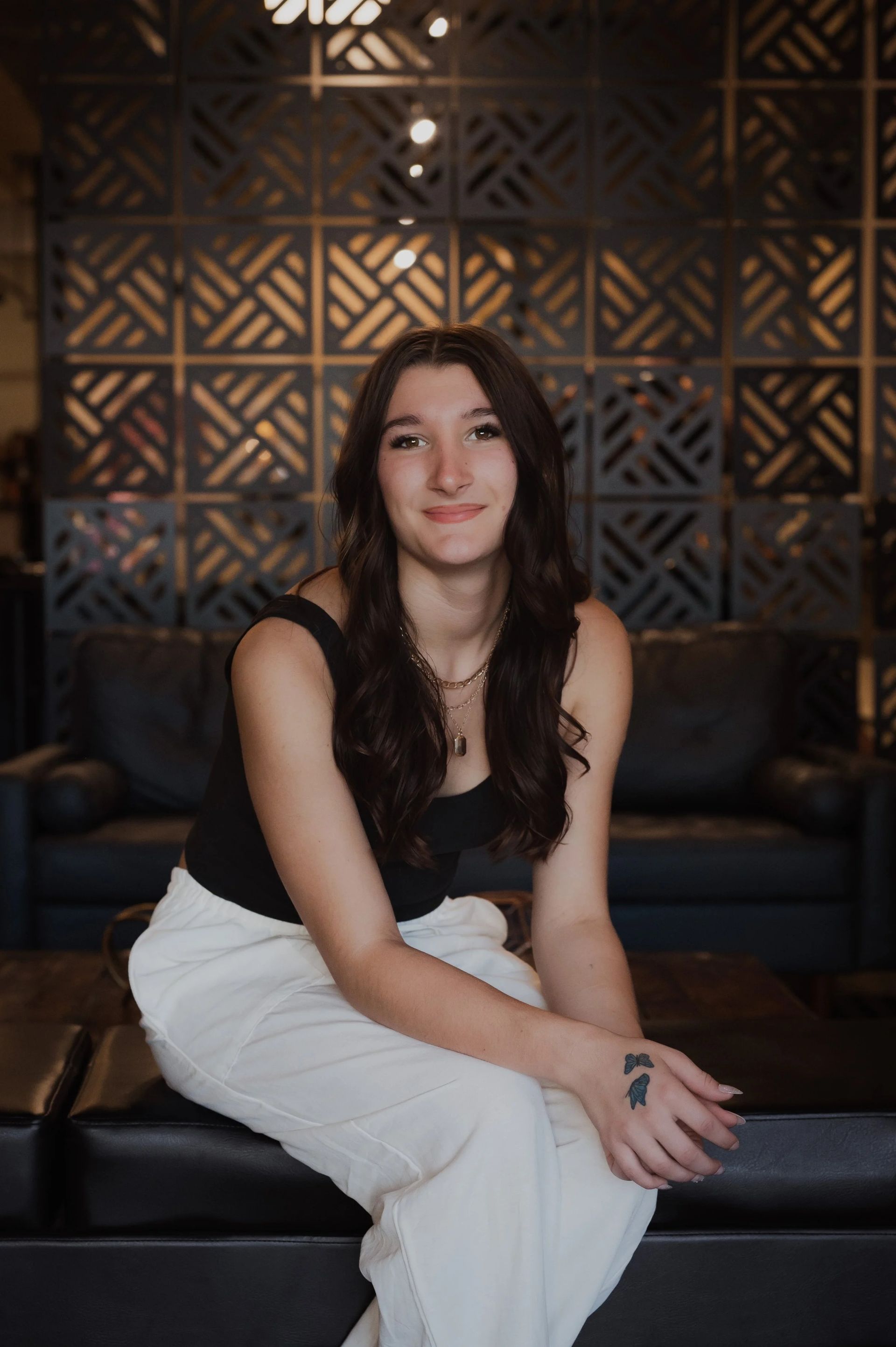 Woman in black top and white pants sits on a black bench, smiling. Dark patterned wall in background.