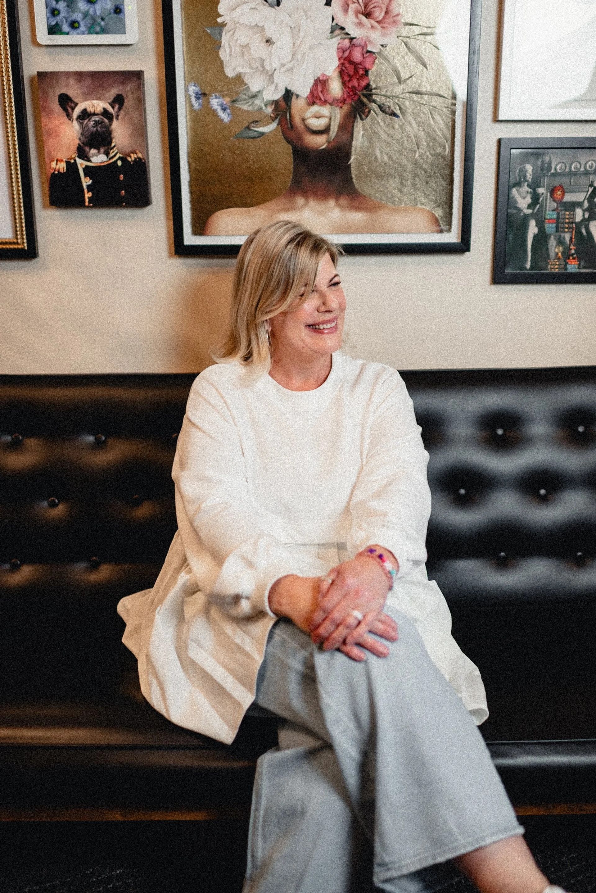 Woman sitting on a black bench, smiling, wearing white sweater and gray pants. Gallery wall in background.