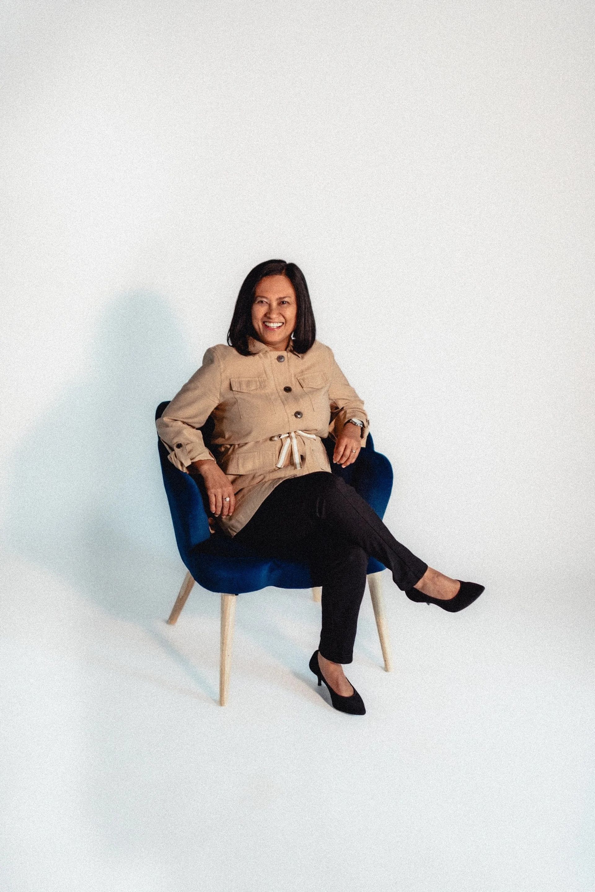 Woman seated in a blue chair, smiling, wearing a tan jacket, black pants, and black shoes, against a white backdrop.