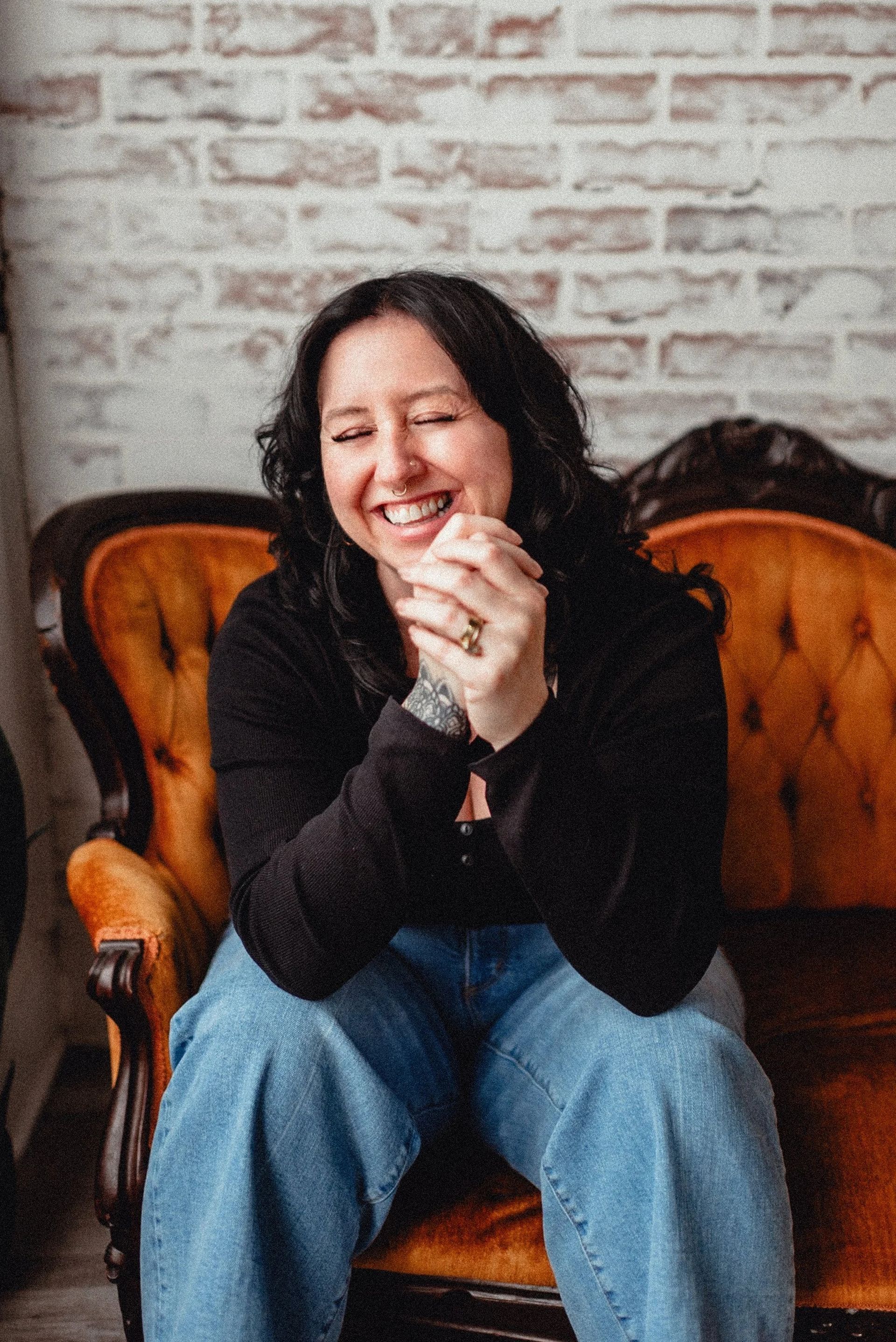 Woman laughing, sitting on a vintage orange couch, hands clasped, in front of a brick wall.