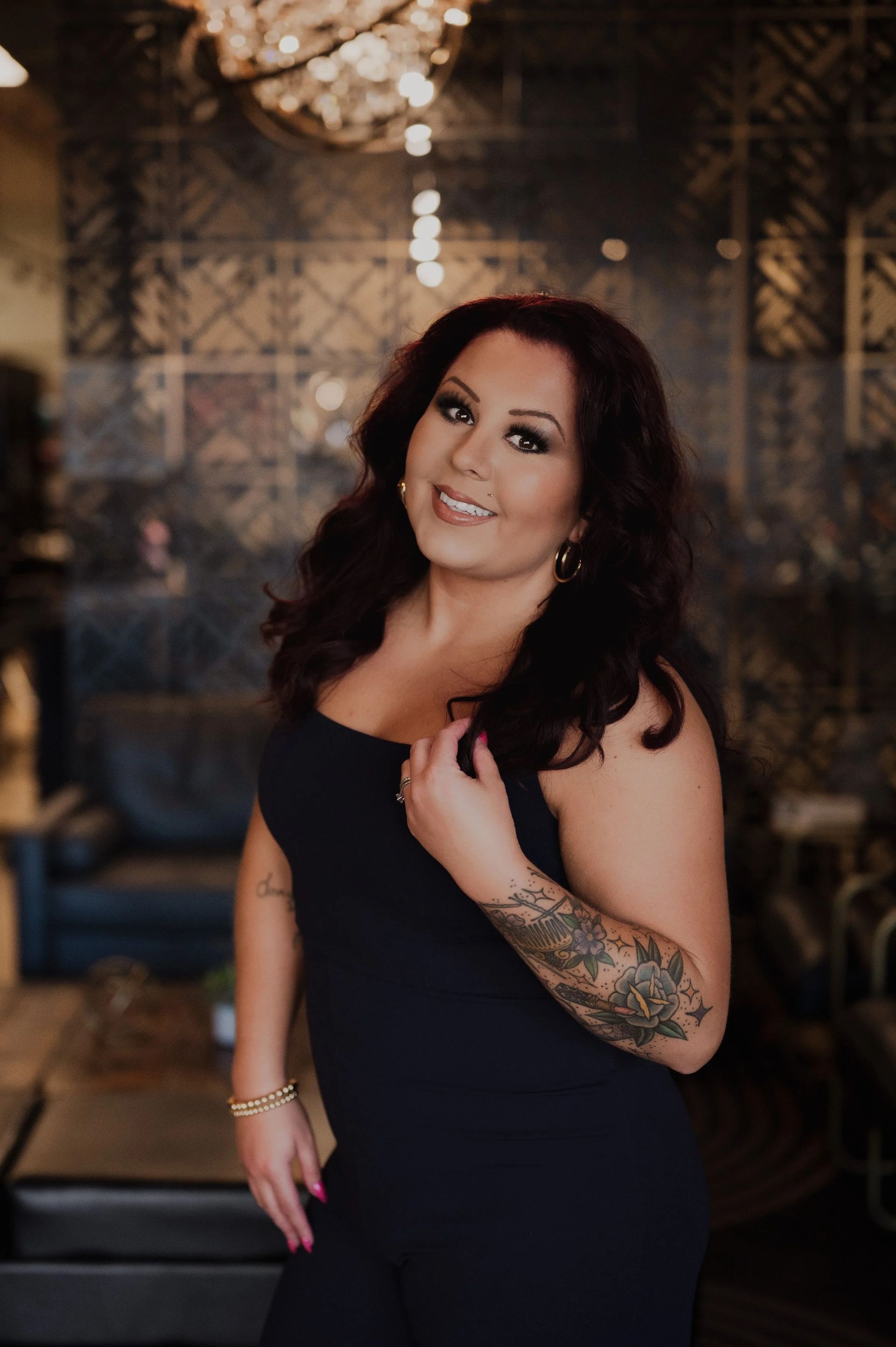Woman with dark hair smiles, stands indoors, wearing a black jumpsuit, with a tattooed arm.