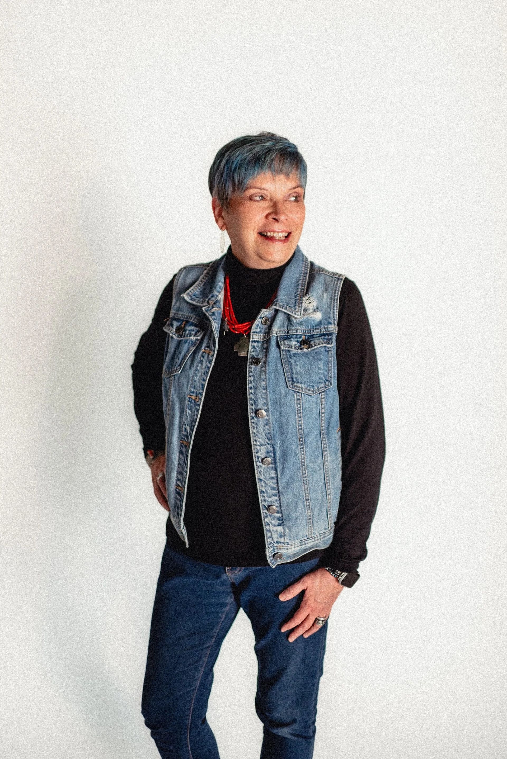 Person in denim vest, black shirt, and jeans smiles, hands on hips, looking right. White background.