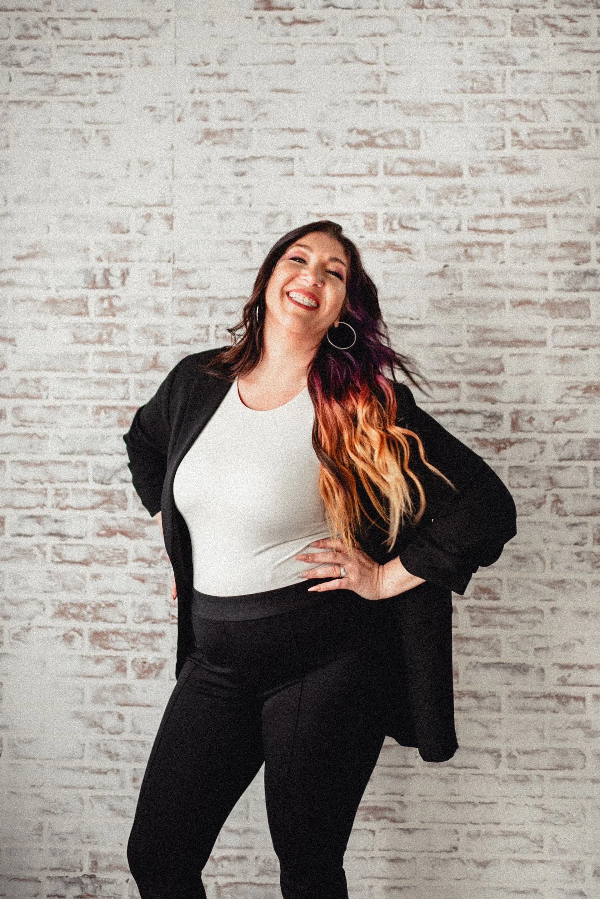 Woman smiles, poses in front of a white brick wall. She wears a white top, black pants, and a black jacket. Hair is highlighted.