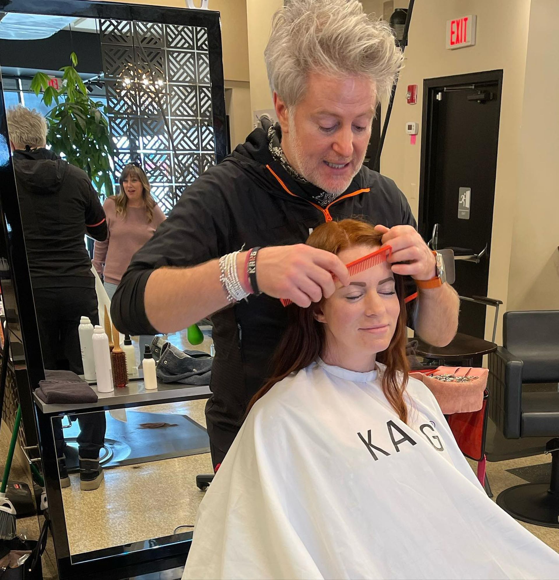 Hair stylist using a comb on a client's red hair in a salon. Another person watches.