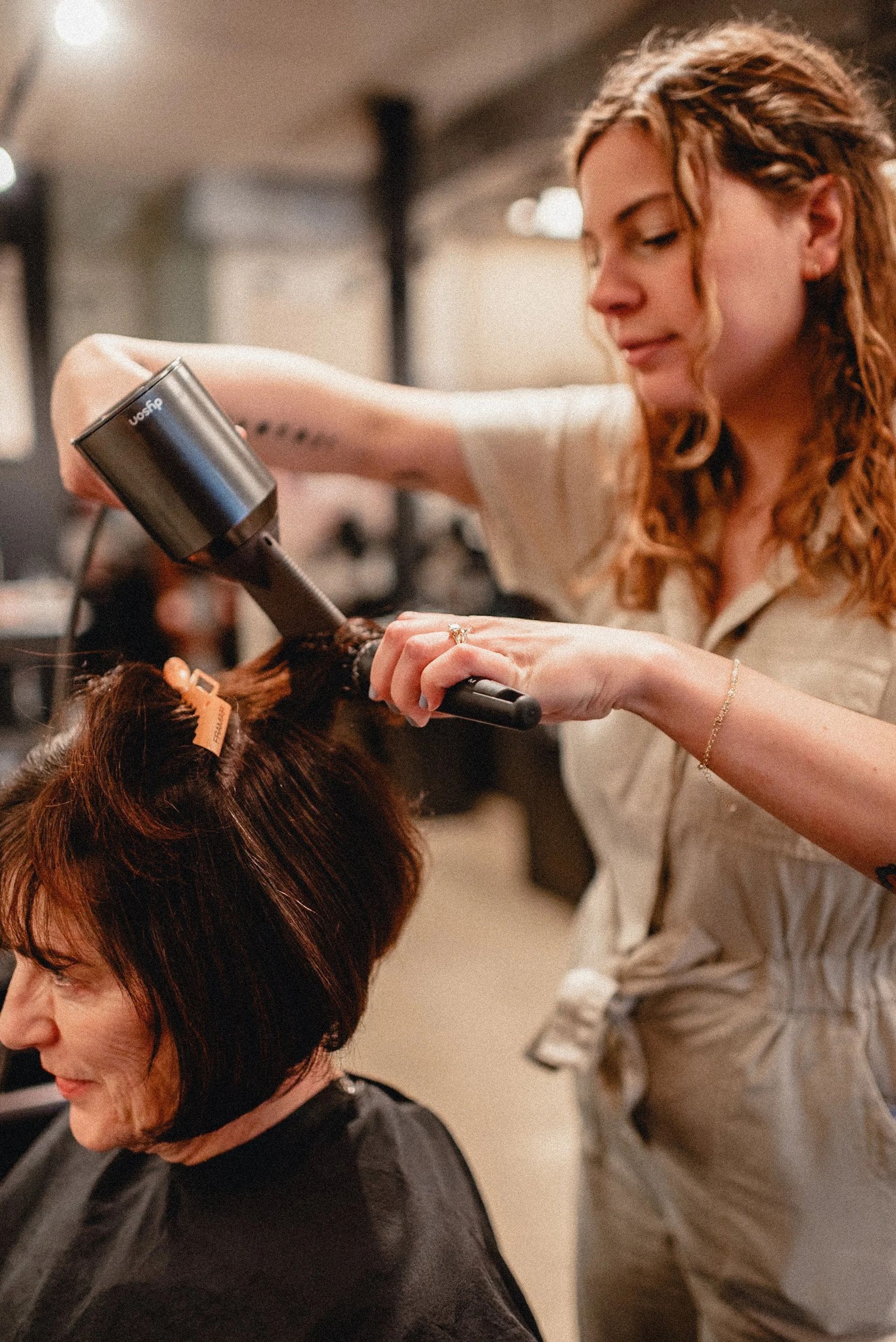 Hairdresser blow-drying a client's hair in a salon. The stylist is using a large blow dryer.