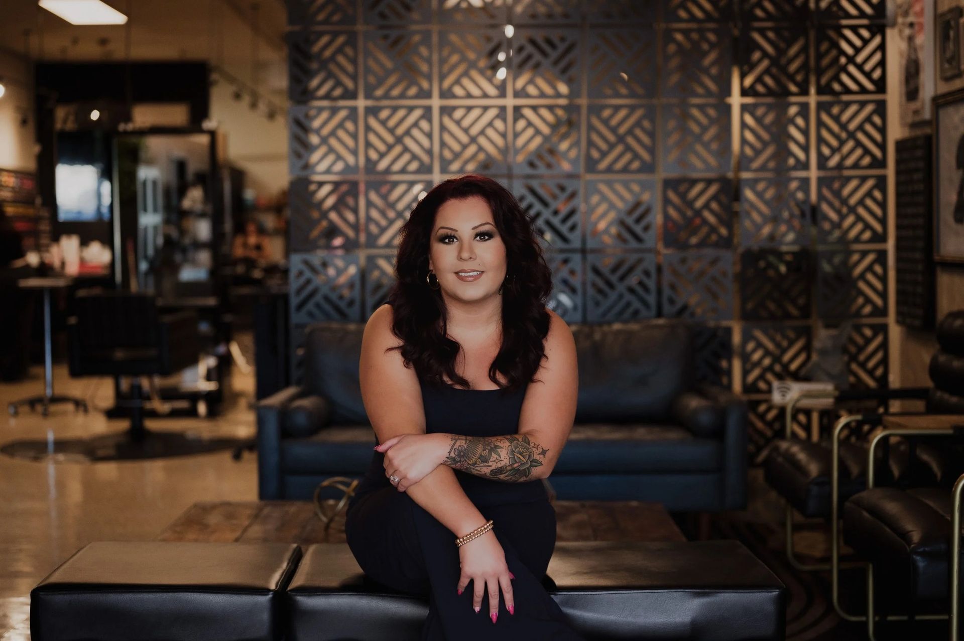 Woman with arm tattoos sits on a dark bench, smiling in a salon.